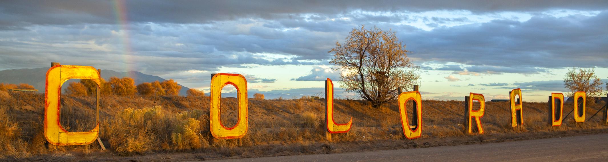 A picture of latters spelling out Colorado with a rainbow in the background on a road near Alamosa