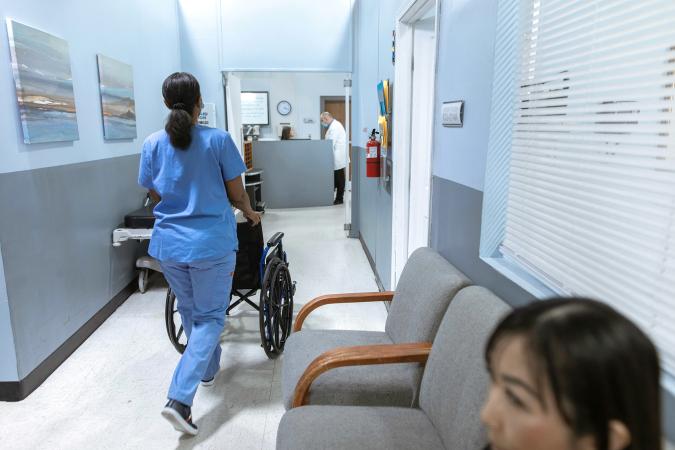 A nurse pushes an empty wheelchair down a hallway as a woman sits in a chair waiting for a doctor