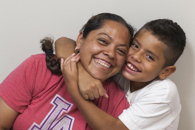 A mom in a red T-shirt smiling and leaning to the right toward her eight-year-old son as he smiles and wraps his arms around her neck