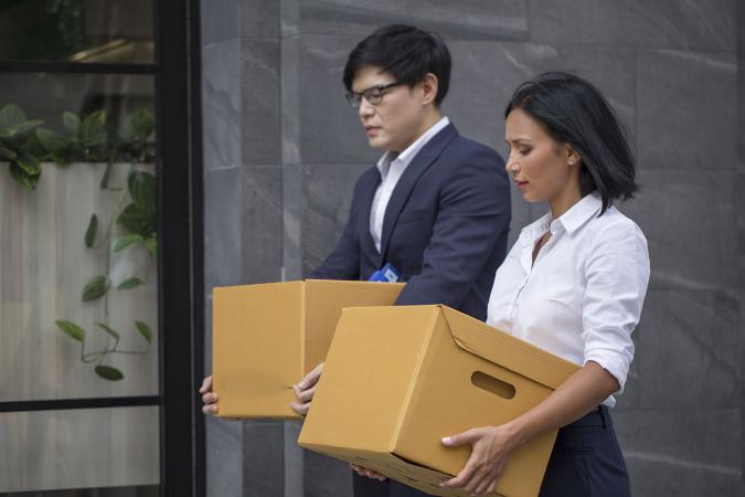 A man in a gray suit and a woman in a white blouse carry cardboard boxes while walking next to an office building