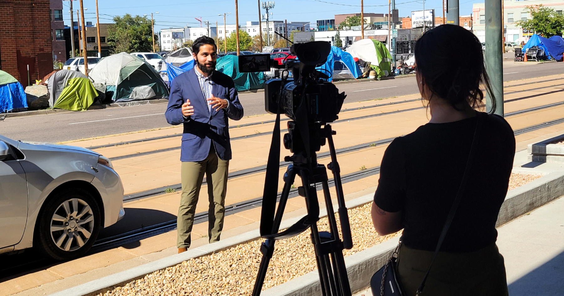 Karam Ahmad being filmed by a KUSA reporter with a homeless encampment in the background
