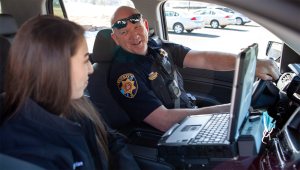 A male police officer talks to a female mental health professional in the front seat of a police car.